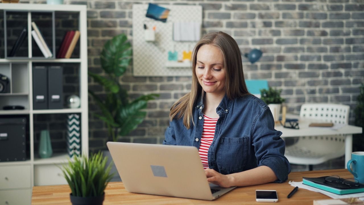 Smiling woman working on a laptop in a contemporary office setting with a brick wall backdrop.